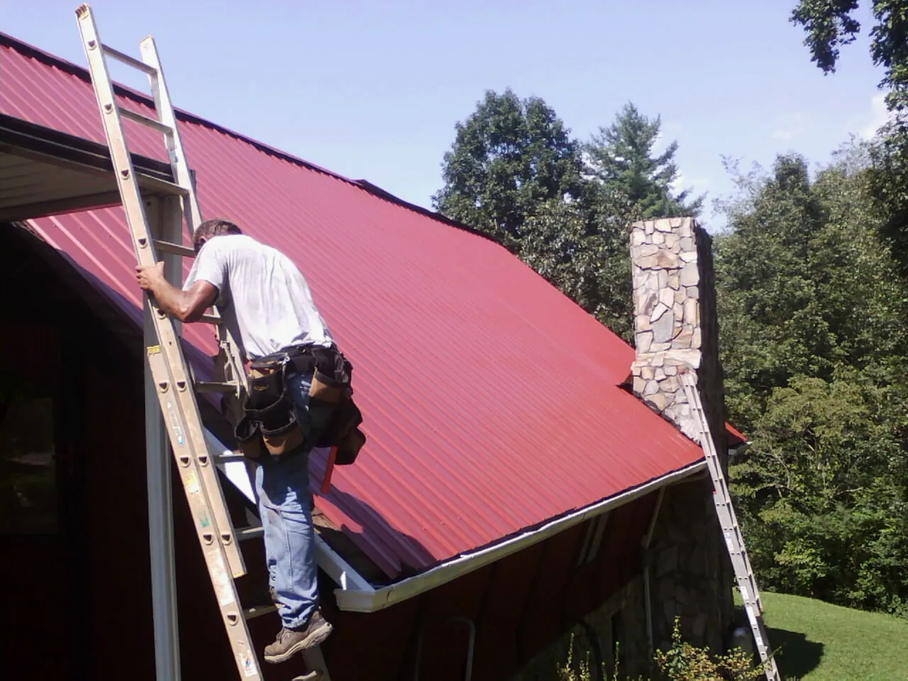 Roofer on a ladder beside a red metal roof and stone chimney — Soffit & Fascia Repair by Roofing Brothers in Otter Creek